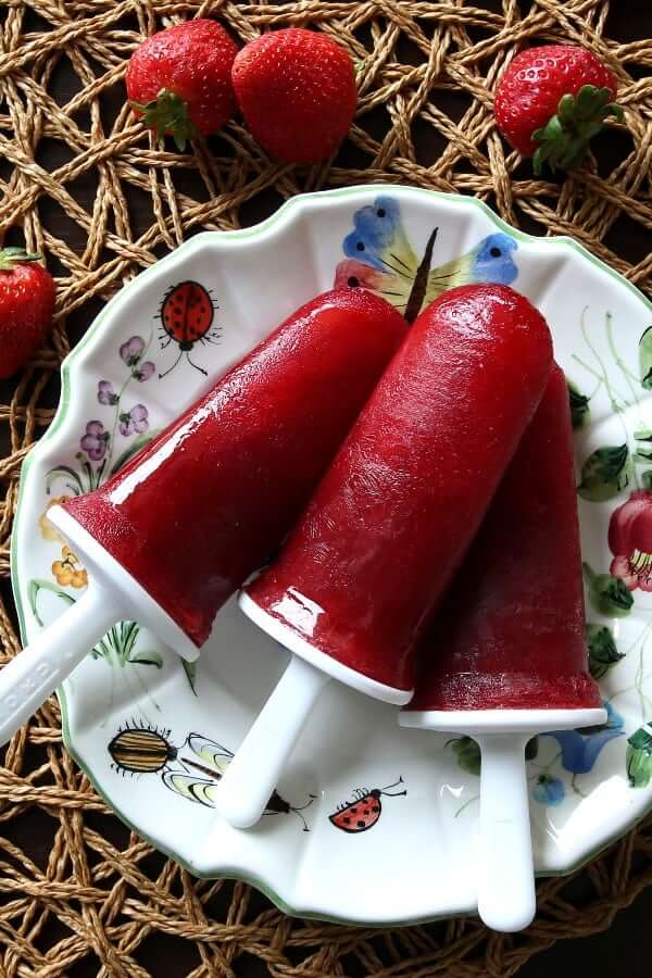 Overhead view of three pomegranate strawberry popsicles popsicles on a flowered plate and woven mat.