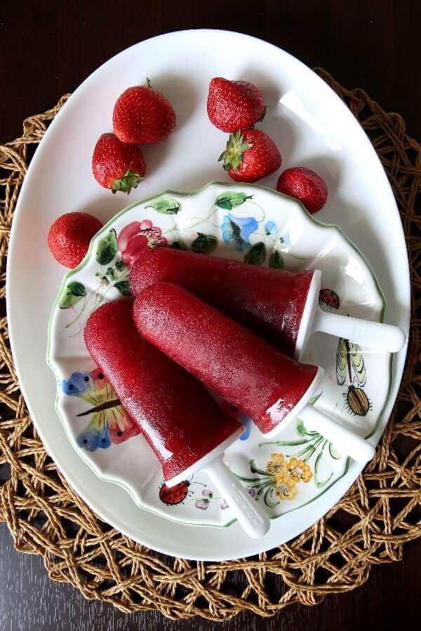 Overhead view of three popsicles on a flower plate and white platter with strawberries.