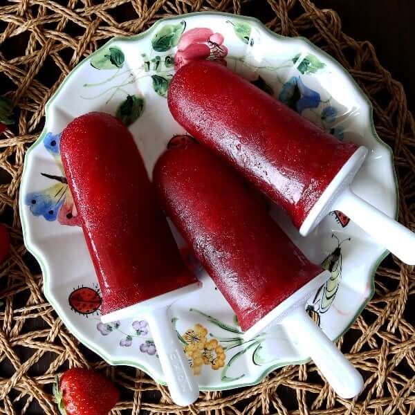 Overhead photo of three pomegranate strawberry popsicles on a flowered plate with strawberries around.