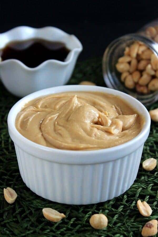 Straight view of peanut butter in a white bowl on an forest green mat with peanuts around.