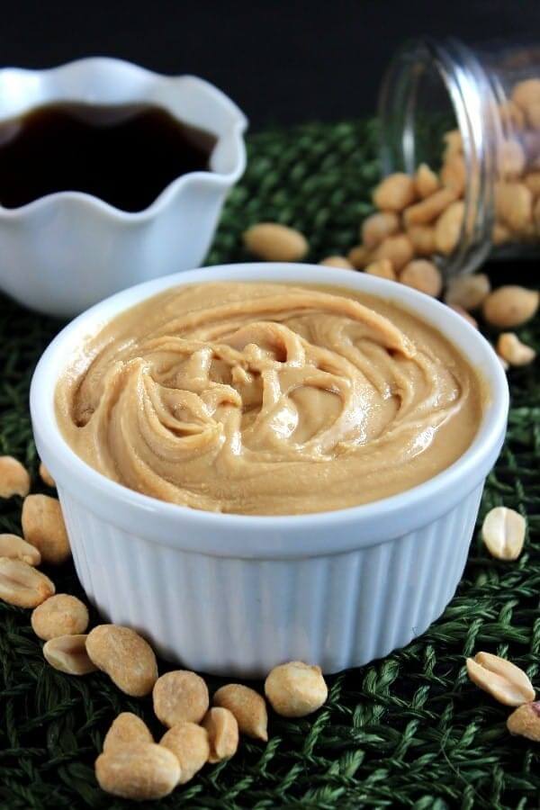 Creamy peanut butter is swirled in a white bowl with peanuts spilled on a green mat below.