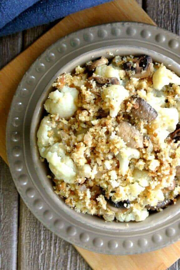 Overhead view of cauliflower and mushrooms with topping in beige bowl with backgrounds of wood and pottery.