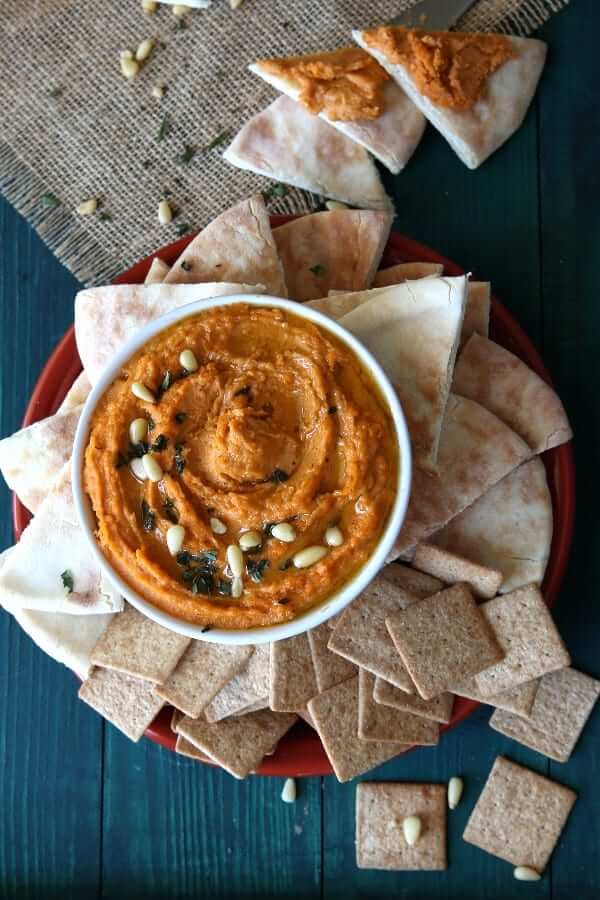 Overhead photo of spicy autumn orange dip on a burnt orange plate surrounded by pita bread triangles and crackers.