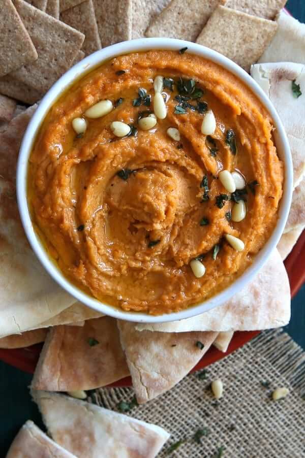 Overhead photo of spicy autumn orange chipotle hummus in a white bowl surrounded by pita bread triangles and crackers.