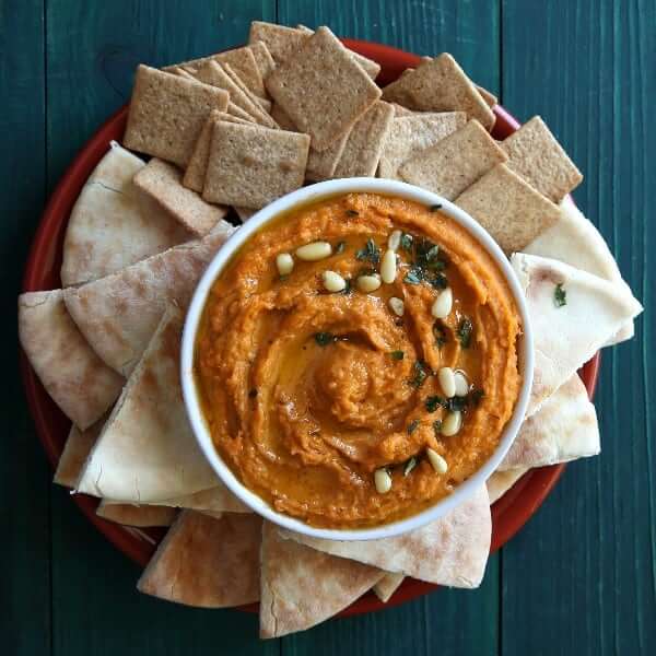 Overhead photo of spicy autumn orange chipotle hummus in a white bowl surrounded by pita bread triangles and crackers.