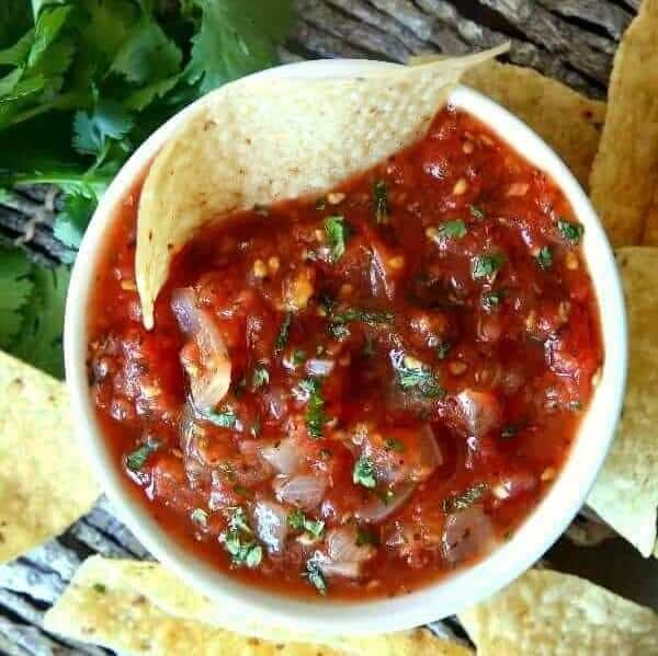 Overhead photo of a white bowl filled with chunky salsa and has tortilla chips scattered around.