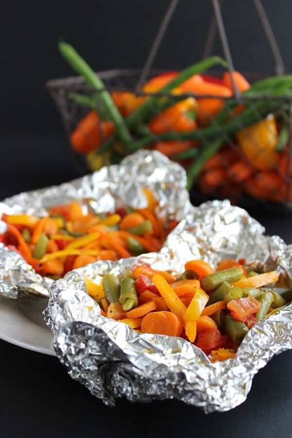 Two opened foil packets showing cooked orange, green and yellow vegetables in front of veggie filled basket.