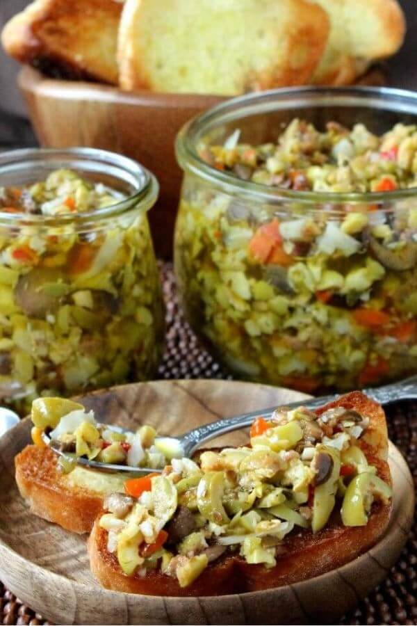 Front view of crostini toast with green tapenade spoon over with jars filled behind.