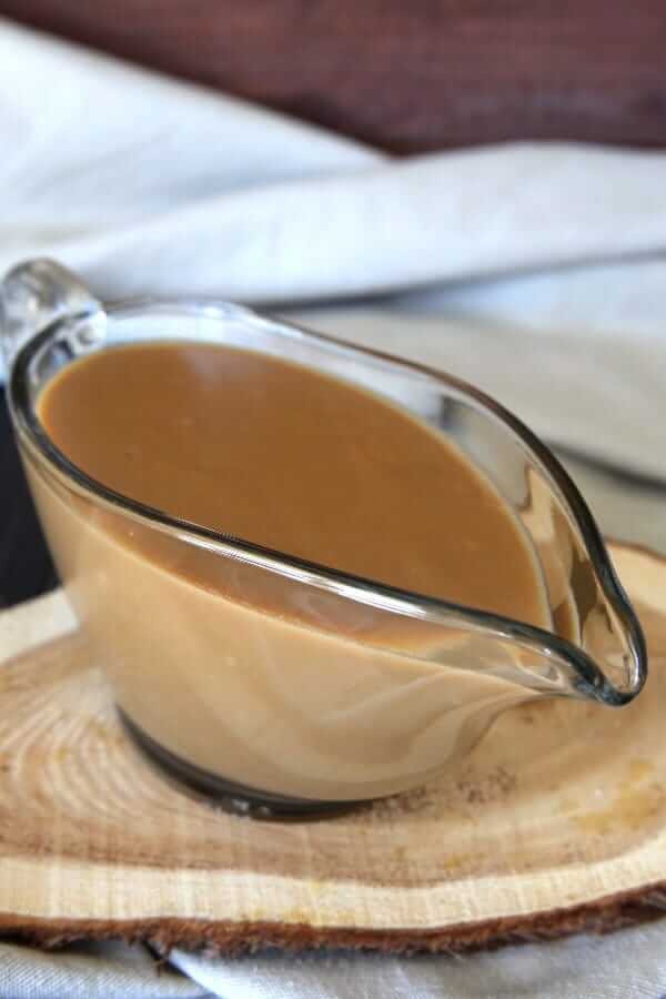 A clear glass gravy boat is filled with Vegan Thanksgiving Dinner and sitting on a wooden trivet.