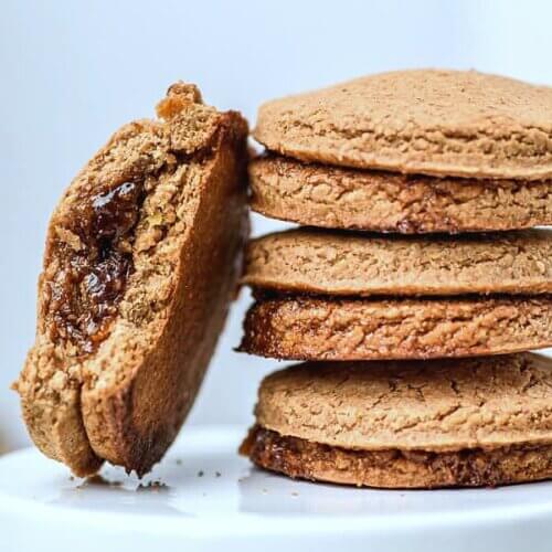 Stack of baked cookies with one leaning on the left with a bite taken out of it to show the jam filling.
