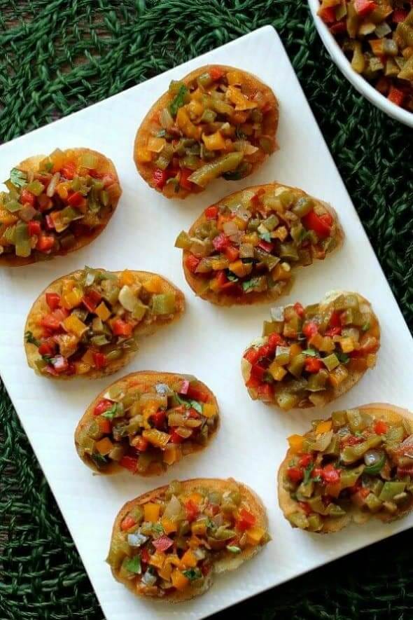 Overhead view of little toasts loaded with colorful diced bell peppers on a rectangular white plate.