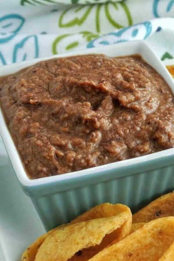 A Black Bean Dip recipe that is filling a turquoise square bowl next to corn chips and a cloth napkin behind.