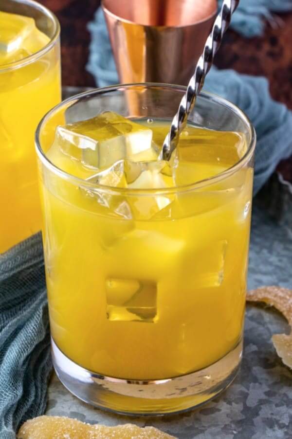 One tumbler filled with an orange drink filled with ice cubes with a metal stirrer inside.