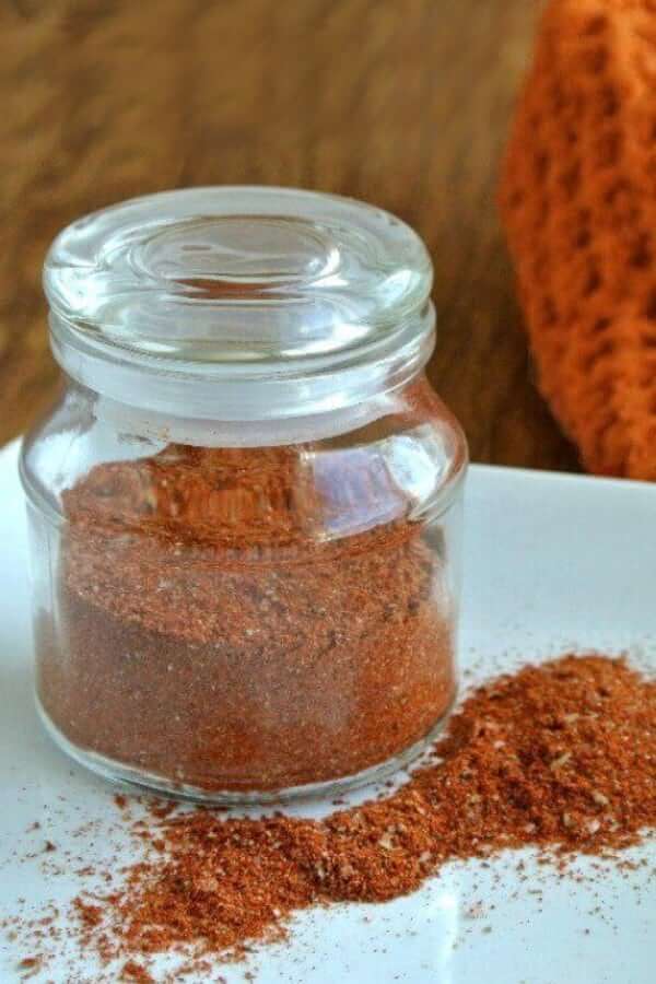 Homemade Taco Seasoning is jarred in a clear glass jar with a spill of rusty taco seasoning color on a white plate.