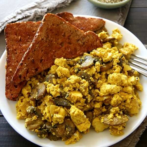 White plate with a large helping of scrambled breakfast with mushrooms and toast and a fork on the side.