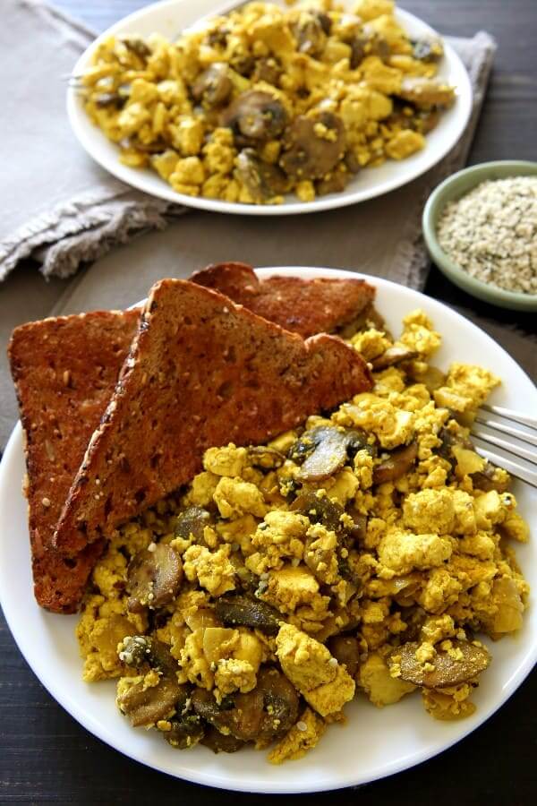 White plate with a large helping of scrambled breakfast with mushrooms and toast on the side.