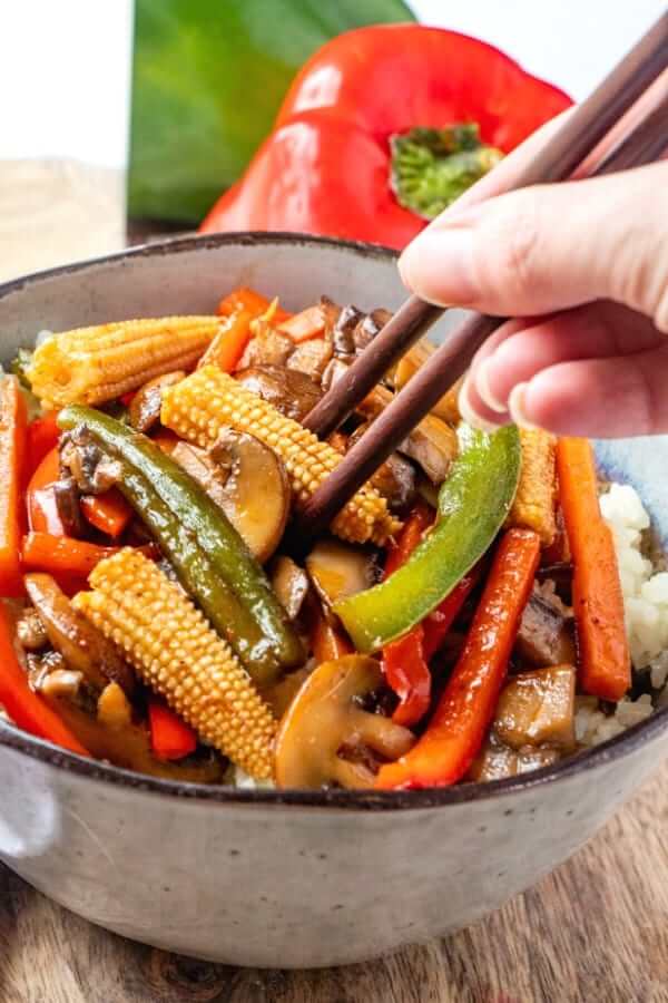 Close up tilted bowl with veggie stir fry over rice and a hand with chopsticks taking a bite.