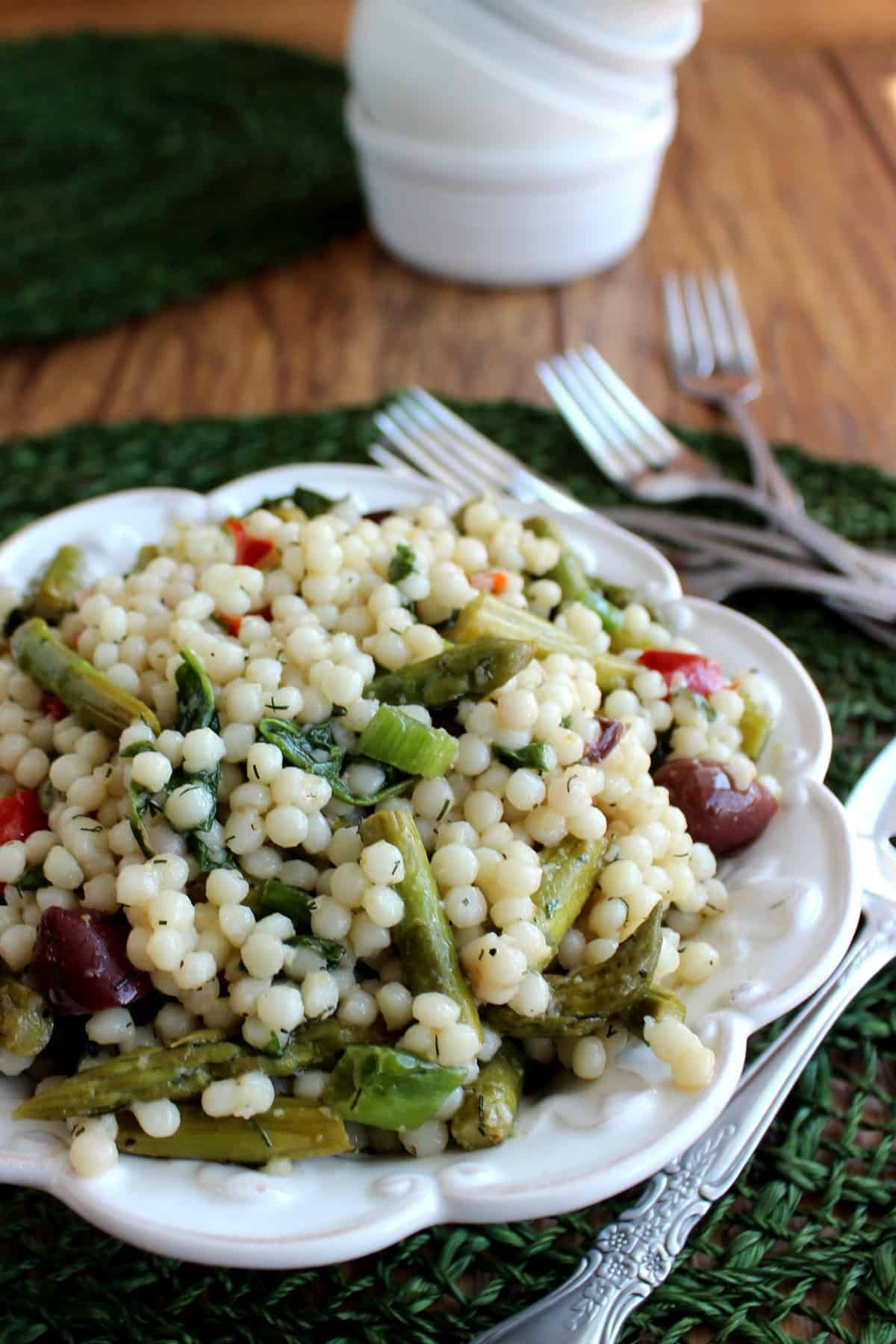 Close-up of a tilted scalloped bowl filled with green vegetables, purple olives and creamy white couscous.