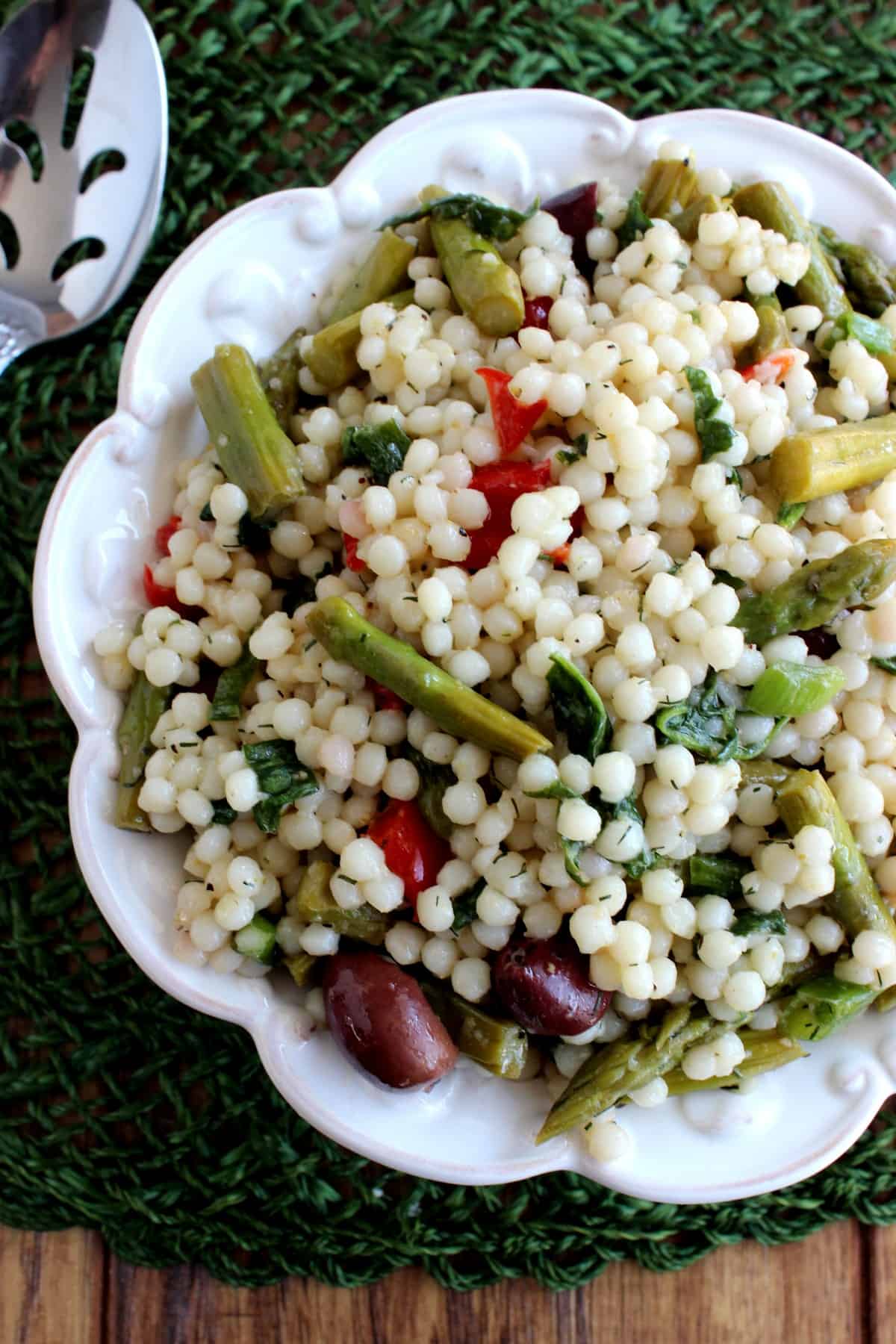 Overhead view of a cold salad in a white bowl filled with couscous, asparagus and more veggies.