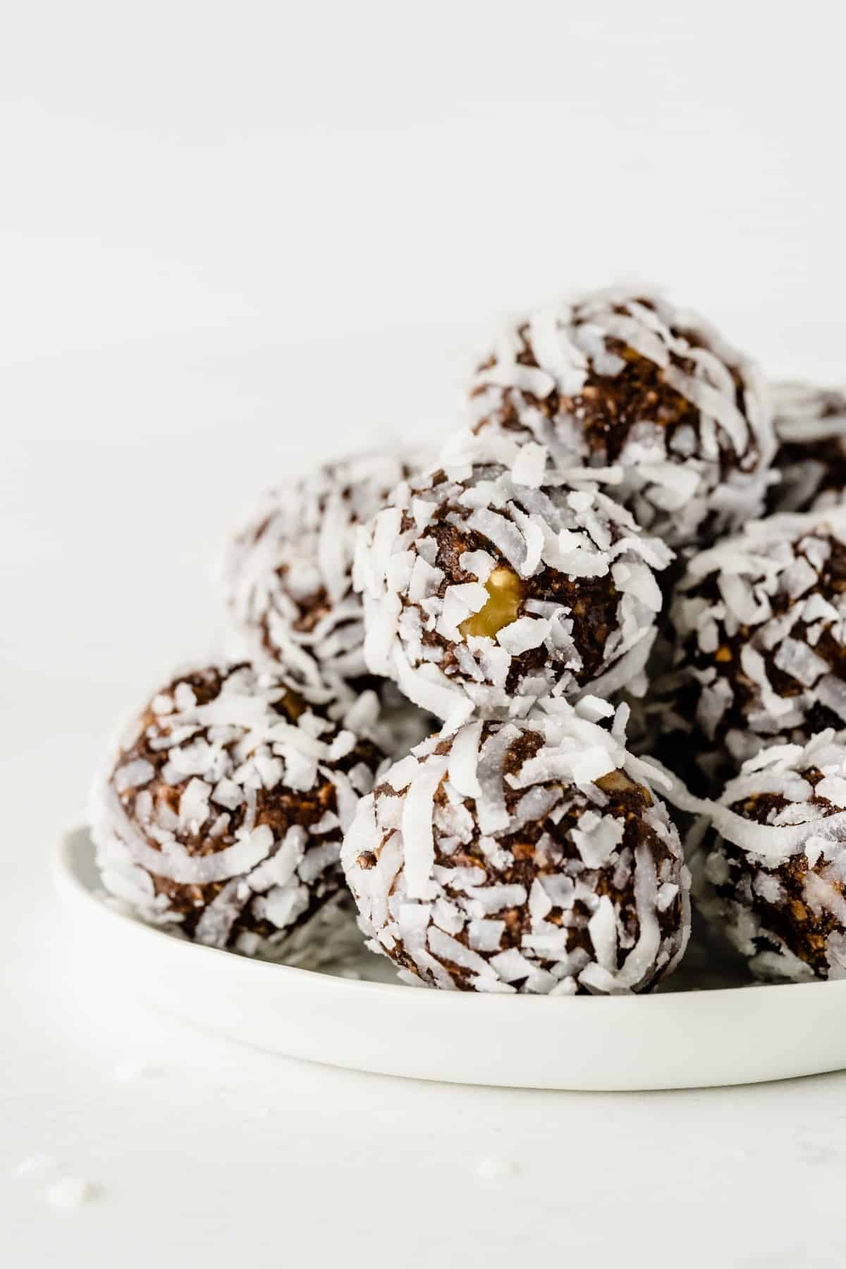 A close-up of chocolate date balls covered in coconut and stacked on a white plate against all white.