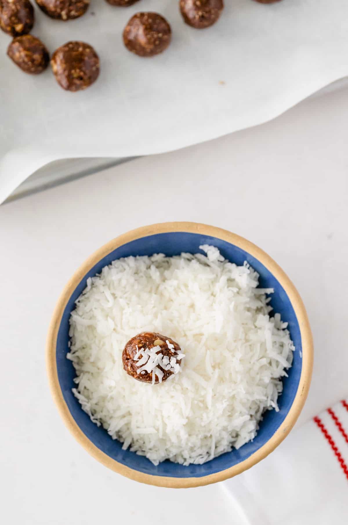 Looking down in a blue bowl full of shredded coconut and one ball is being rolled in the coating.