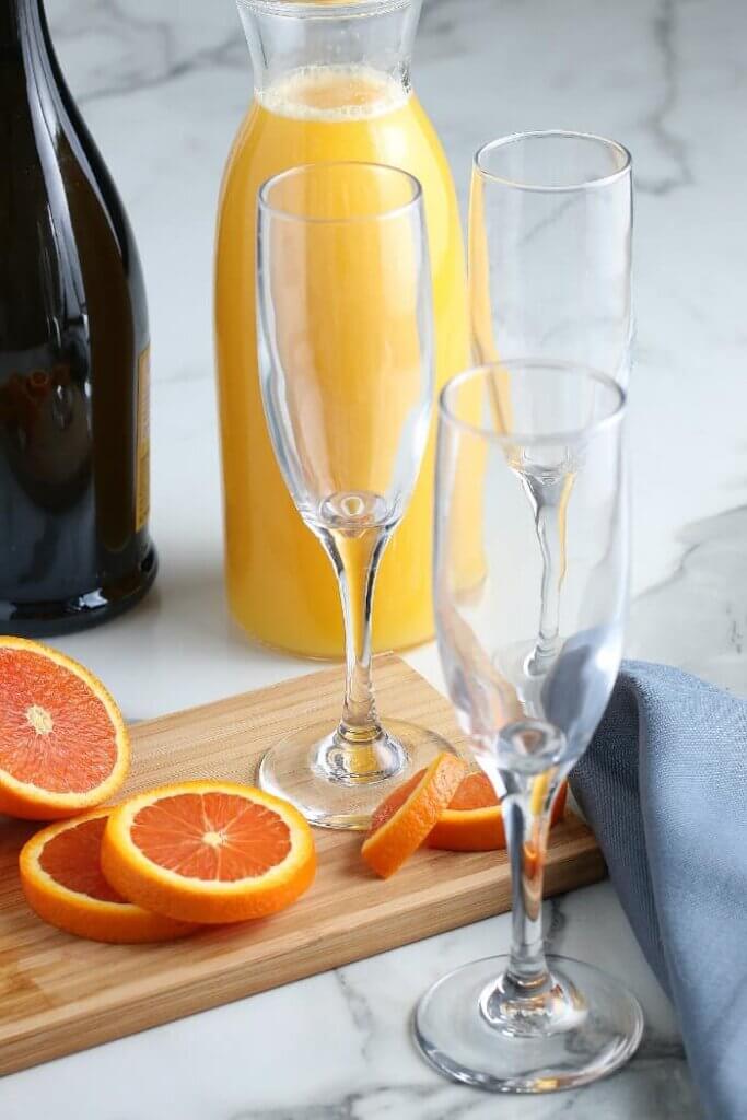 Empty glass set up with fruit garnish on the wooden cutting board and sparkling wine behind.