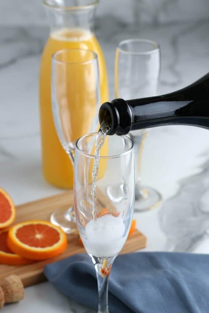 Sparking wine is being slowly poured into an empty champagne glass with slices of oranges behind on a cutting board.