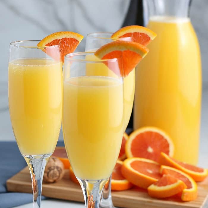 Three filled and garnished champagne filled with golden mimosas with lots of orange slices for garnish behind.