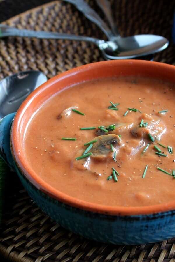 Cropped blue handled bowl filled with thick creamy tomato soup on a straw mat.