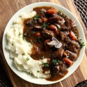 Square photo showing rich beef bourguignon on mashed potatoes and with the plate on a wood grained server.