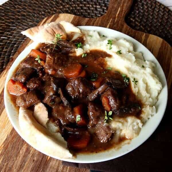 Angled white plate on a wooden cutting board filled with mashed potatoes and a saucy stew.