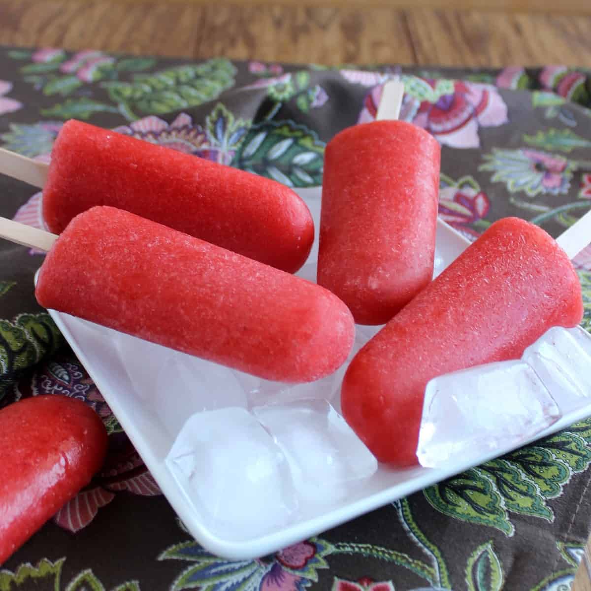 Four apple strawberry popsicles laying on a bed of ice with one laying on the side on a flowered napkin.
