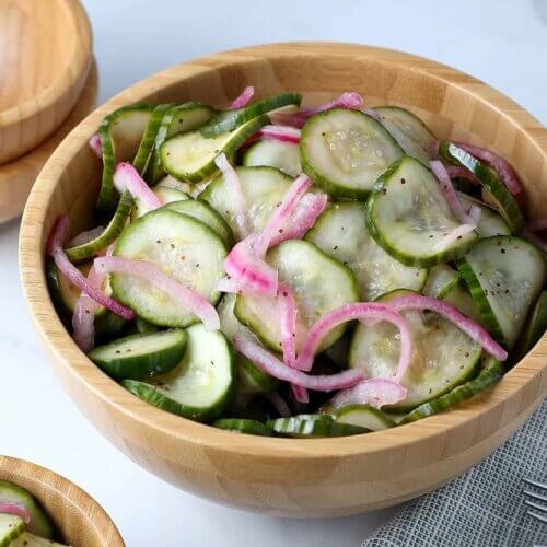 A large wooden bowl is tilted forward and filled with green cucumber slices and thinly sliced red onions.