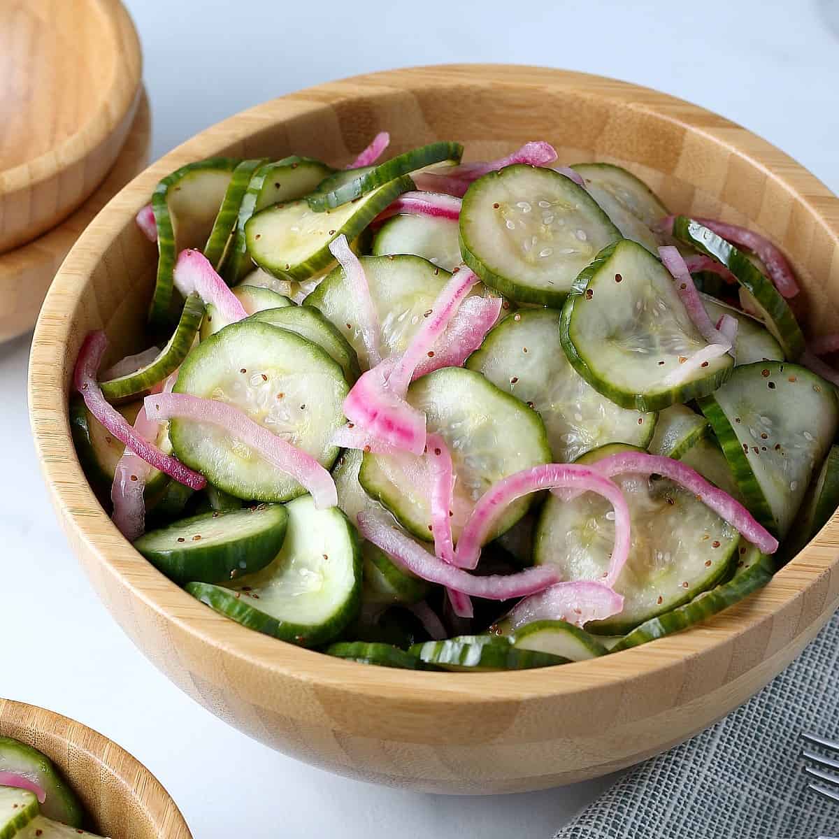 A large wooden bowl is tilted forward and filled with cucumber vinegar salad tharhas green cucumber slices and thinly sliced red onions.