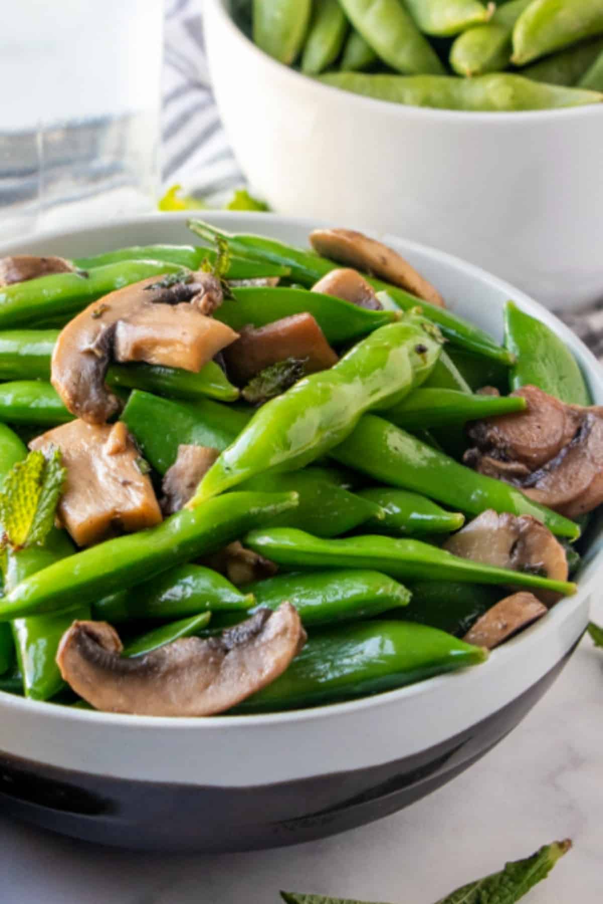 Close up view of glistening a sugar snap peas recipe with mushrooms in a white and silver striped bowl.