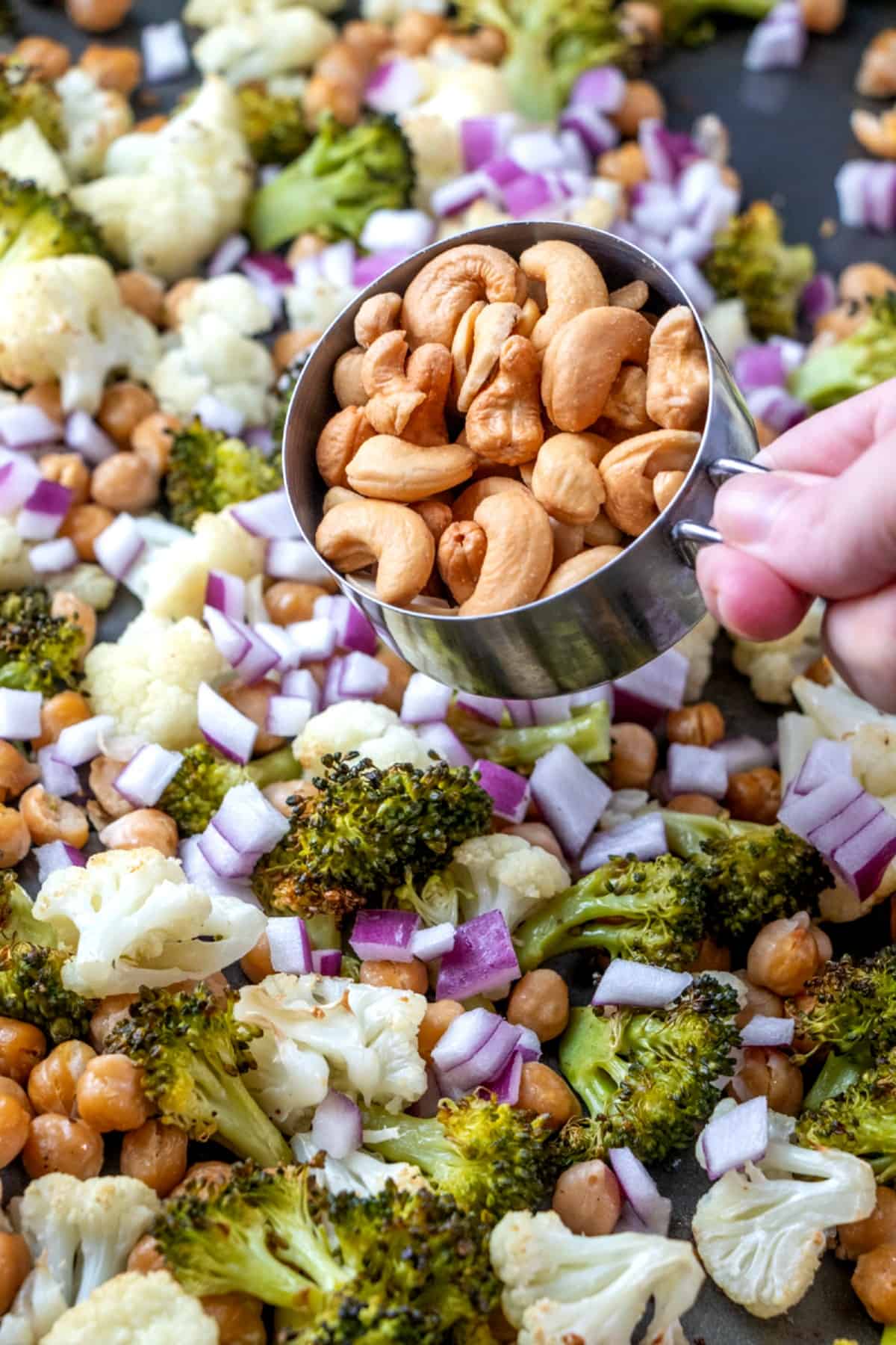 Mixed vegetable bites with cashews are roasted and on a baking sheet fresh diced red onions have been added and now cashews are being sprinkled over all.