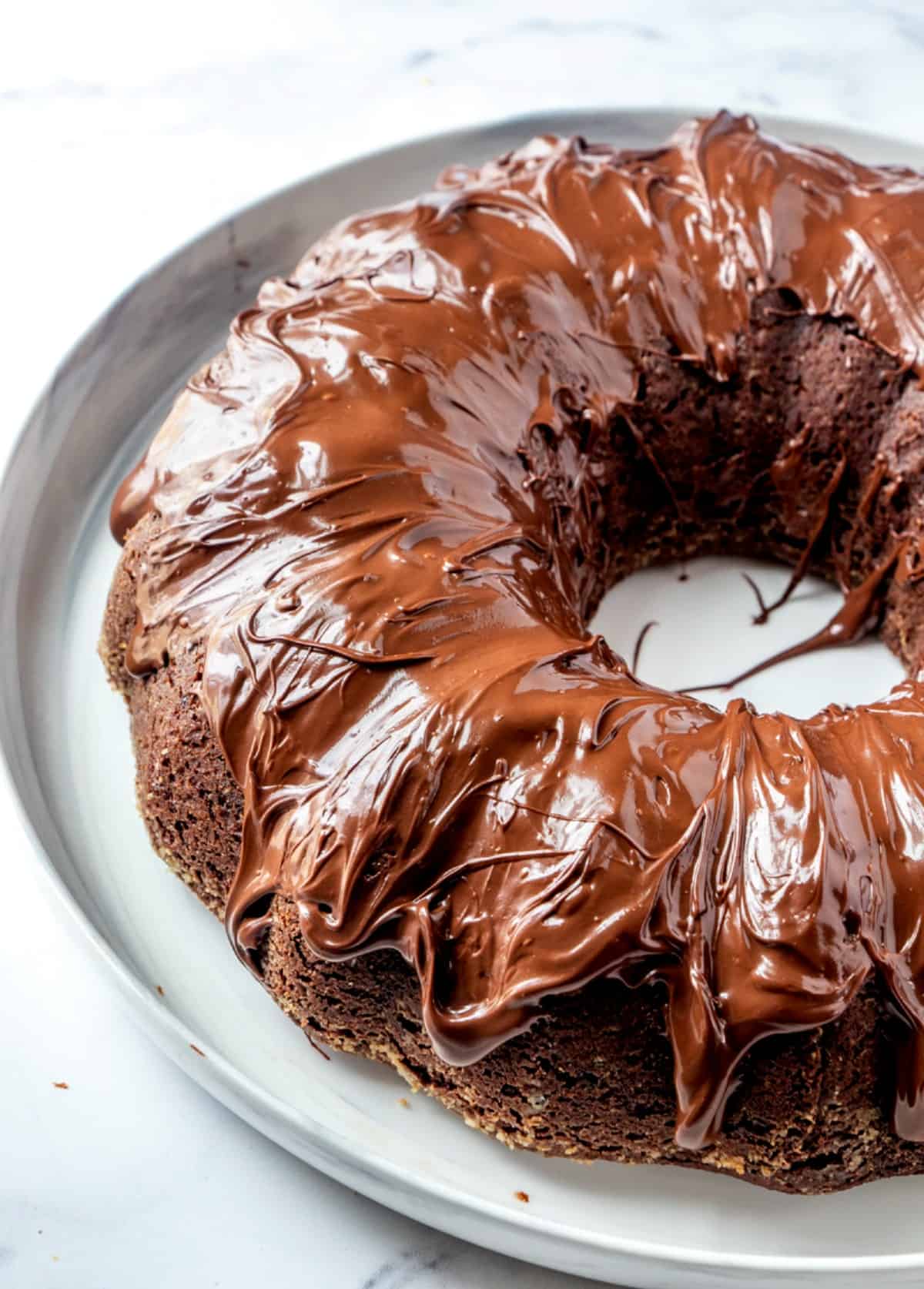 Tilted vegan chocolate bundt cake covered with thick chocolate frosting and on a white plate.