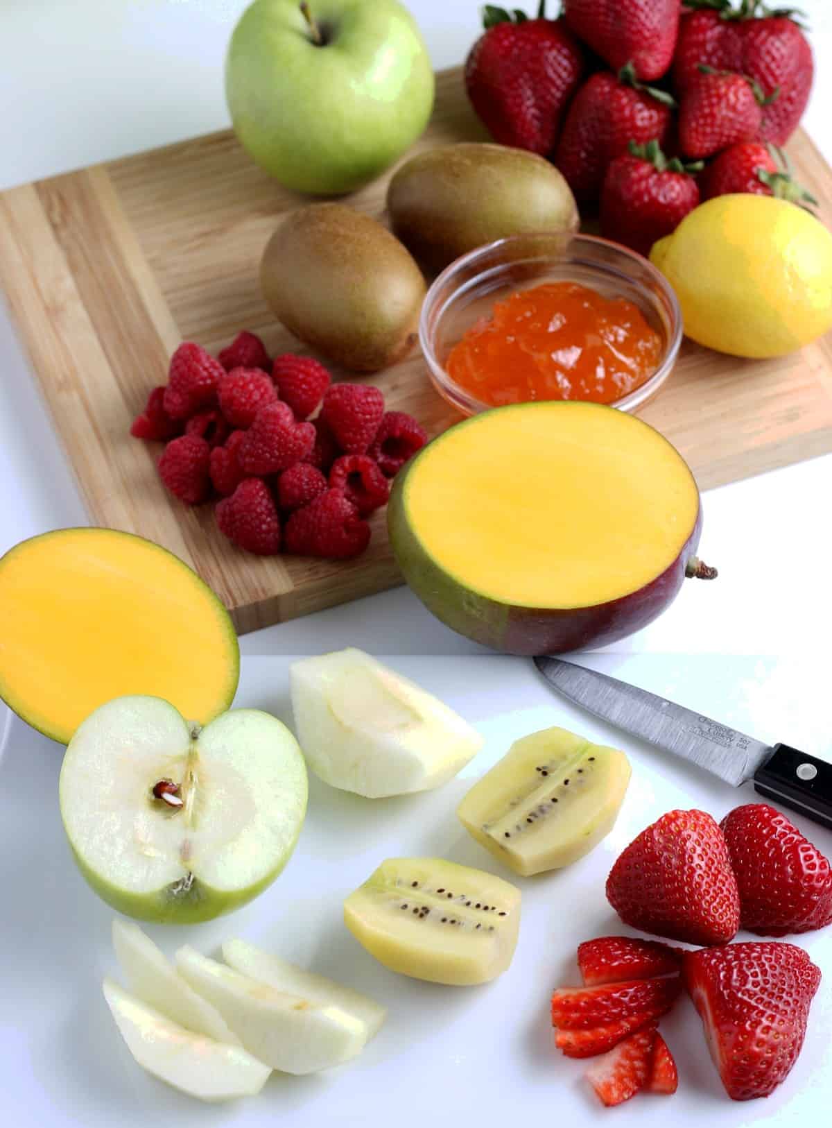 Fruit varieties being diced and sliced on a wooden cutting board with some halves in front.