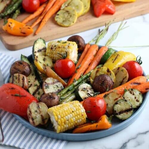 Close angled enamelware platter filled with grilled vegetables with more behind on a wooden board.