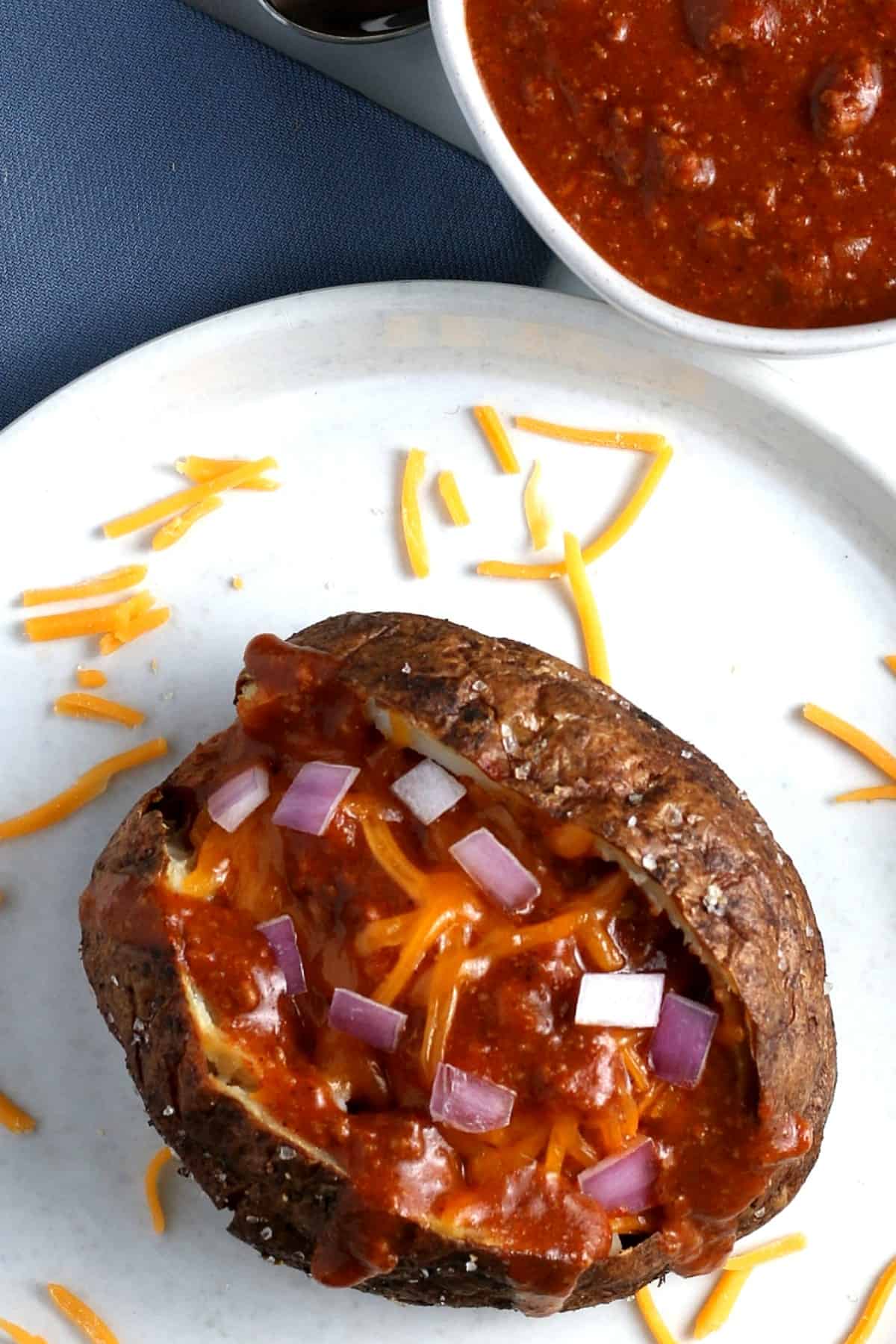Overhead view of a baked potato filled with chili on a white plate with a bowl of serving chili on the side.