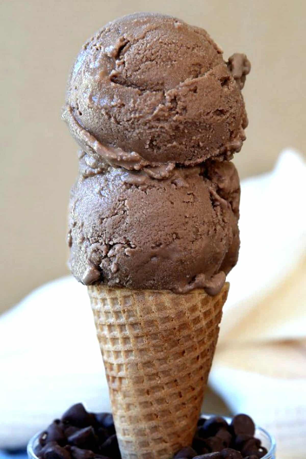 Two scoops of homemade chocolate ice cream on a cone and standing in a bowl of chocolate chips.