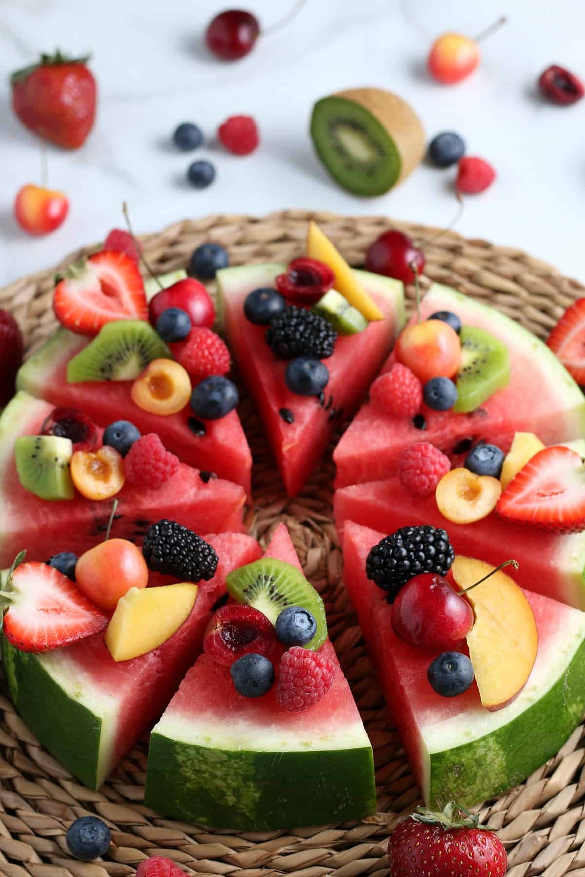 Angled thick slices of watermelon covered with fresh fruit pieces and then sliced into wedges for serving. All on a woven mat.