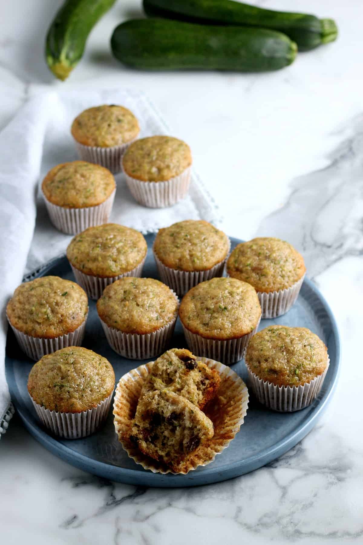 Twelve zucchini muffins on a tray and a white marble counter with the front muffin opened to see the moist insides.
