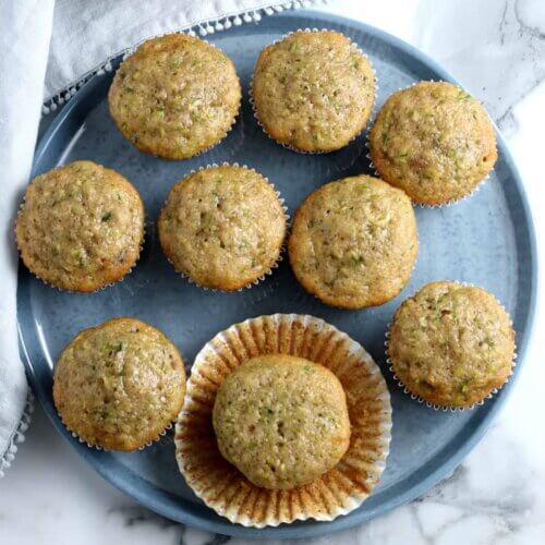 Overhead view of zucchini muffins on silverblue tray where one muffin has it's paper cup opened around it.