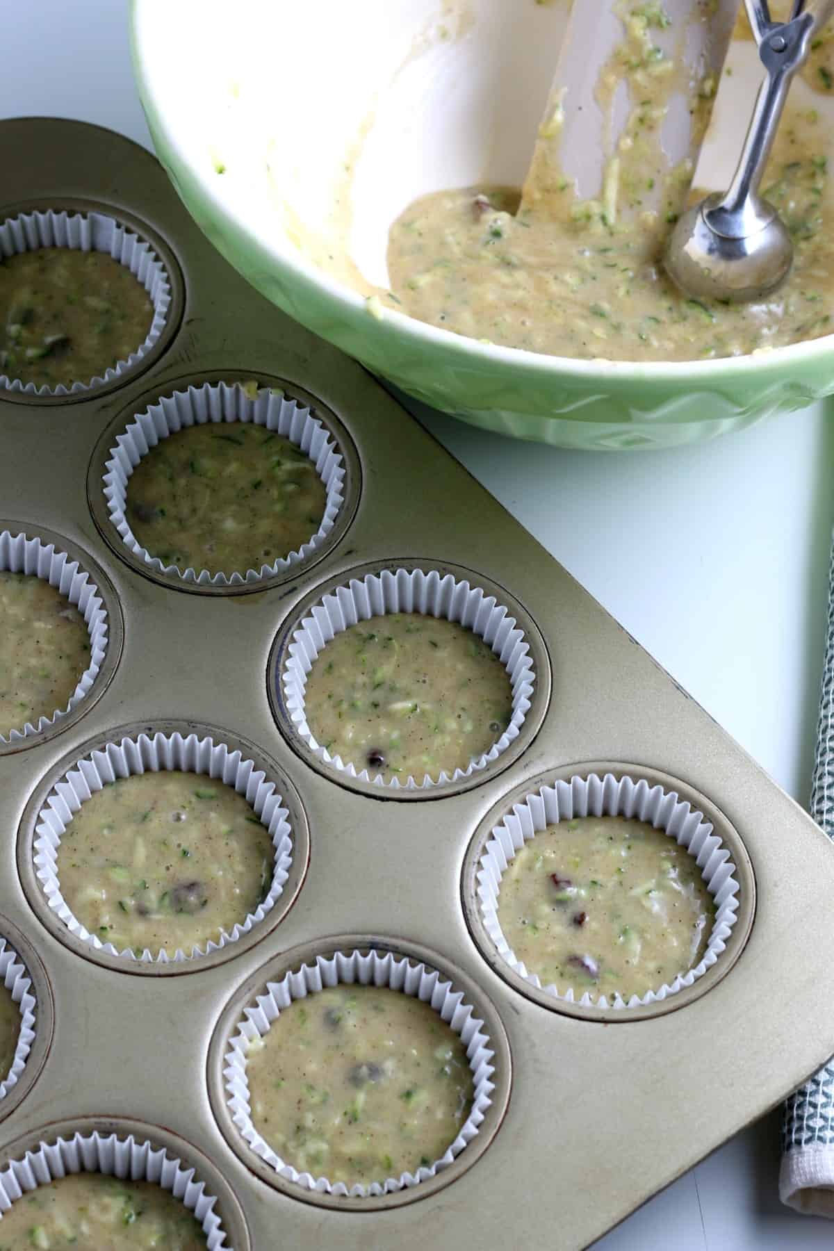 Overhead view of muffin cups in a tin filled with muffin battger. A bowl is on the side with more batter and a scoop.