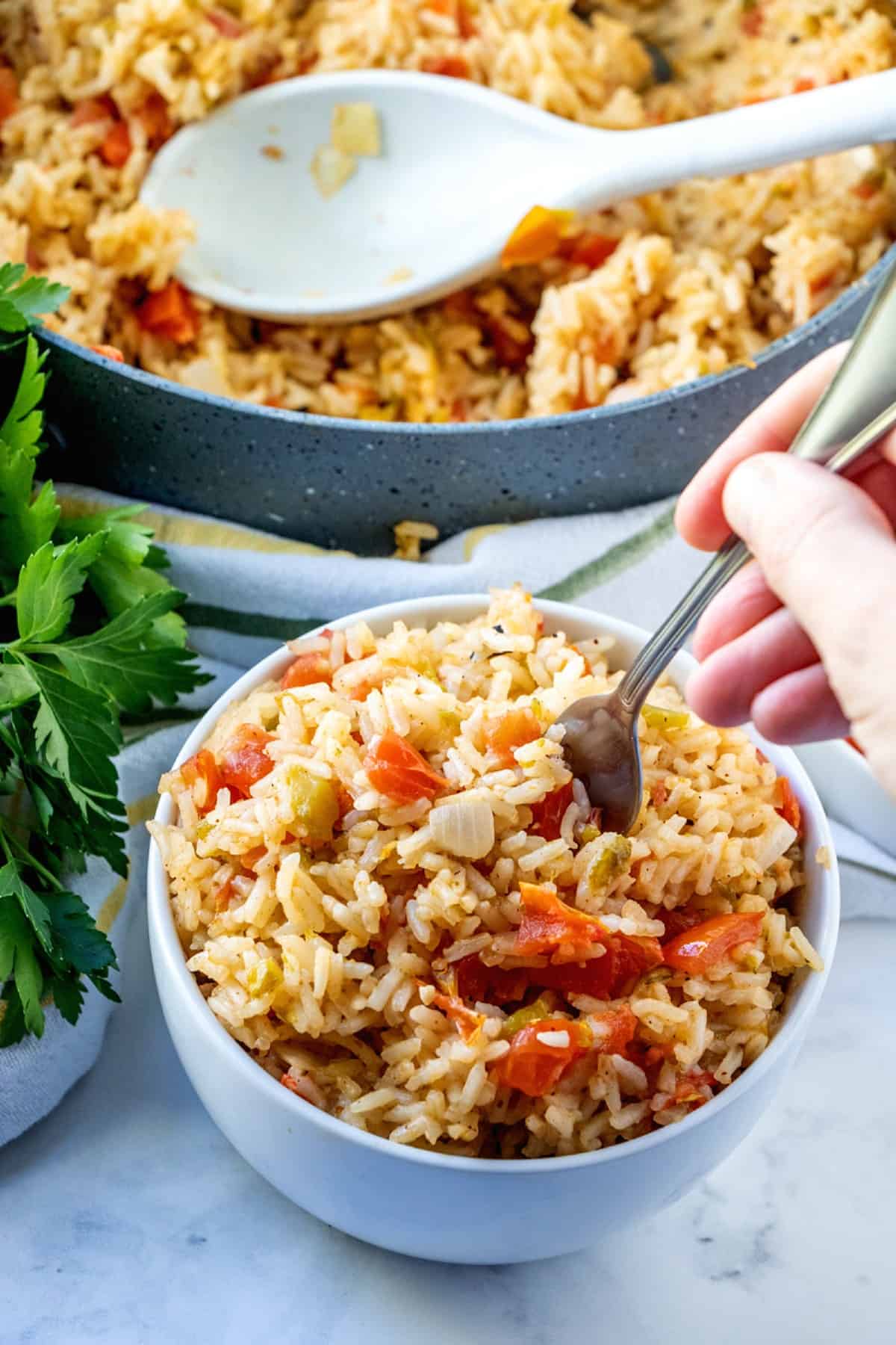 Spanish rice is in a white bowl with a hand poking in a fork for their first bite. The partially full skillet is behind.