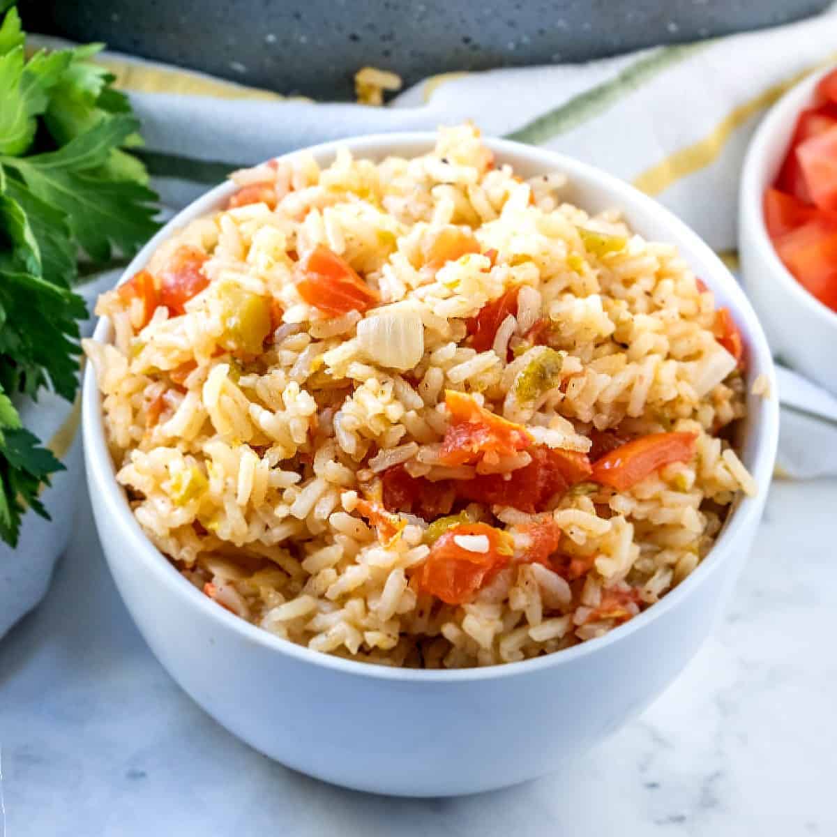 Spanish rice is in a white bowl and is showing off the canned green chilies and tomato pieces. Fresh parsley is on the side.