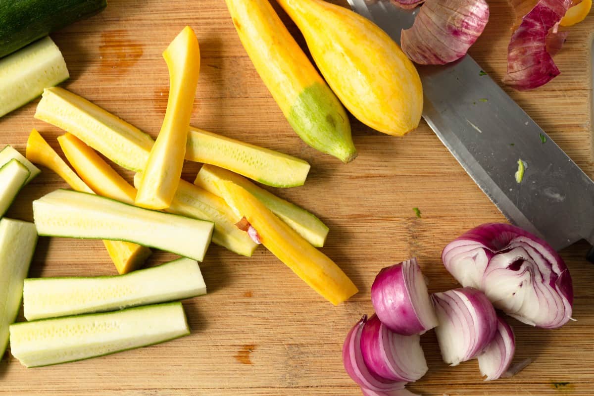 Summer squash, zucchini and red onion are beinf sliced and diced on a wooden cutting board.