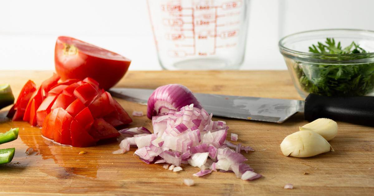 Wide photo showing diced tomatoes and red onion on a cutting board with a knife on it's side.