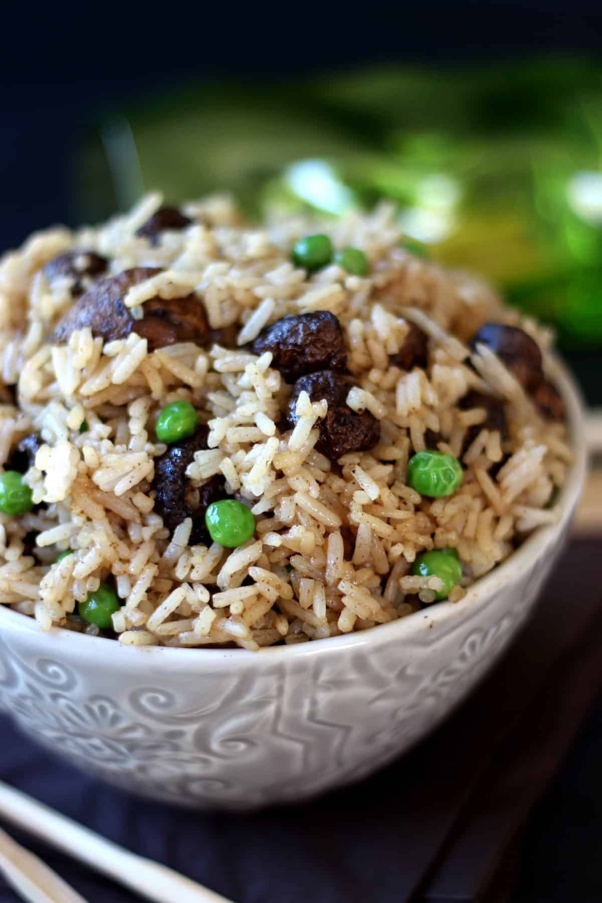 Gorgeous rice with mushrooms and peas is filling a patterned bowl and is tilted towards the camera. A green glass is glimmering in the back.